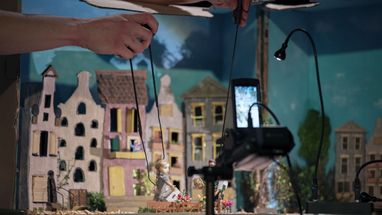 Twee handen spelen een scène in een miniatuurlandschap. Op de voorgrond staat een camera die het tafereel filmt.