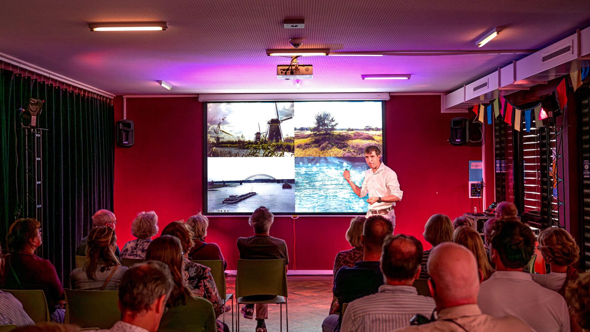 Een spreker presenteert aan een publiek voor een grote projector met beelden op de achtergrond.